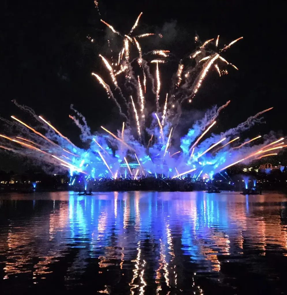 Fireworks display over a dark lake as Disney fireworks cast blue and orange light reflecting on the water. - Favorite Grampy Travels