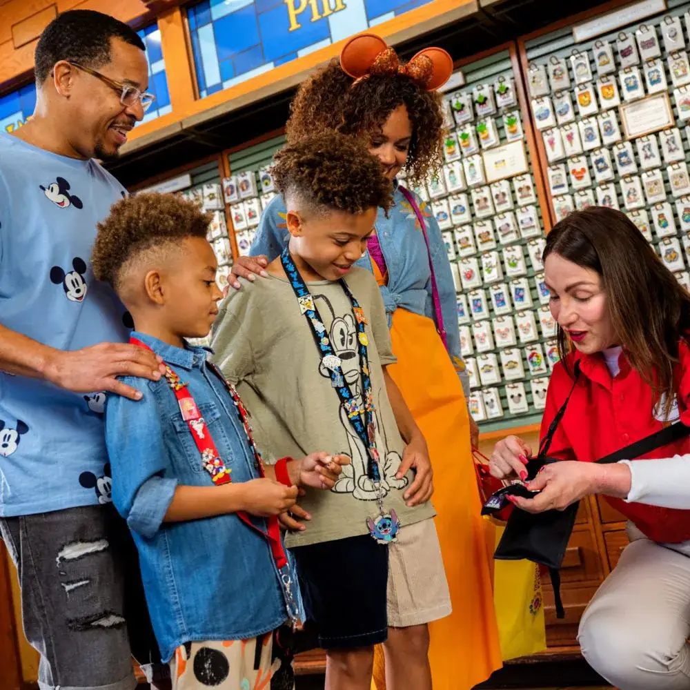 A family at a Disney store watches a cast member in a red polo shirt demonstrate Disney pin trading to the kids. - Favorite Grampy Travels