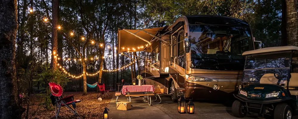 A Class A motorhome parked at Disney’s Fort Wilderness Resort at dusk, featuring a picnic table under an extended awning, string lights, and a golf cart parked nearby. - Favorite Grampy Travels