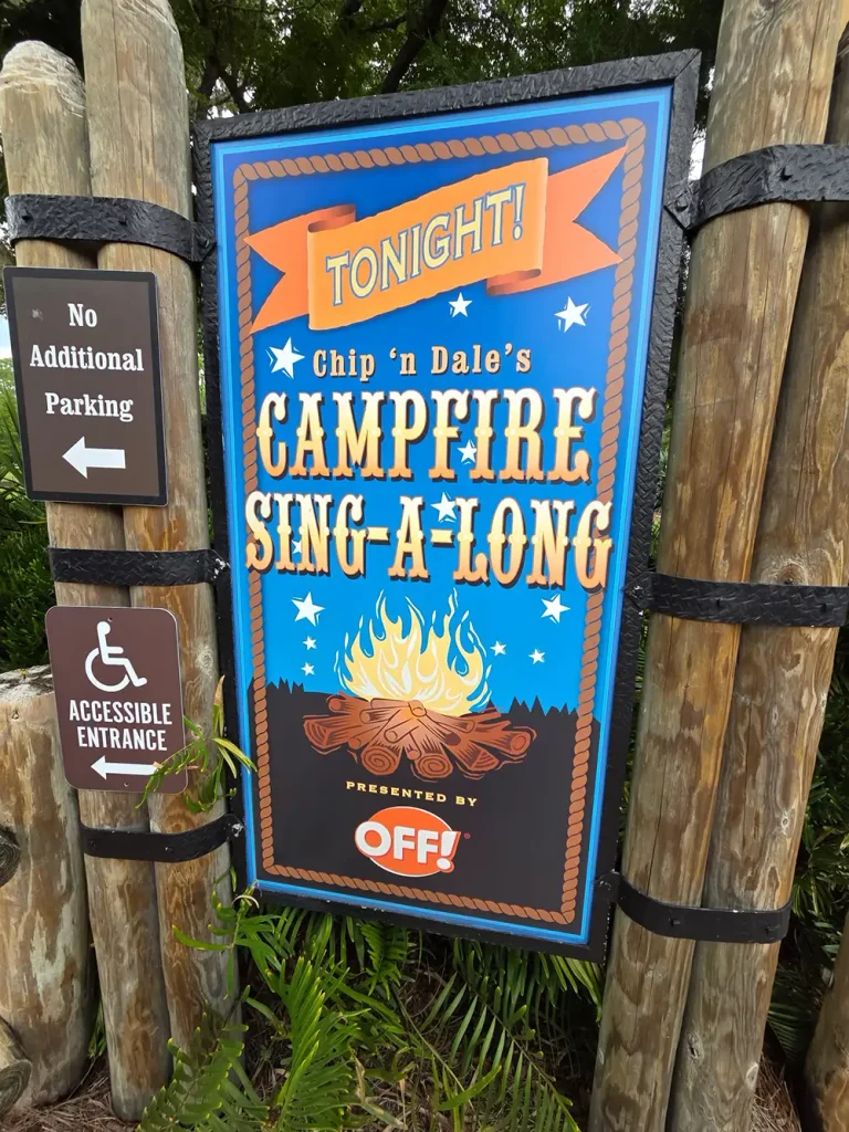 A sign for the Chip ‘n’ Dale’s Campfire Sing-A-Long at Disney Fort Wilderness, showing an illustration of a campfire, with smaller signs for No Additional Parking and Accessible Entrance attached to the wooden posts. - Favorite Grampy Travels