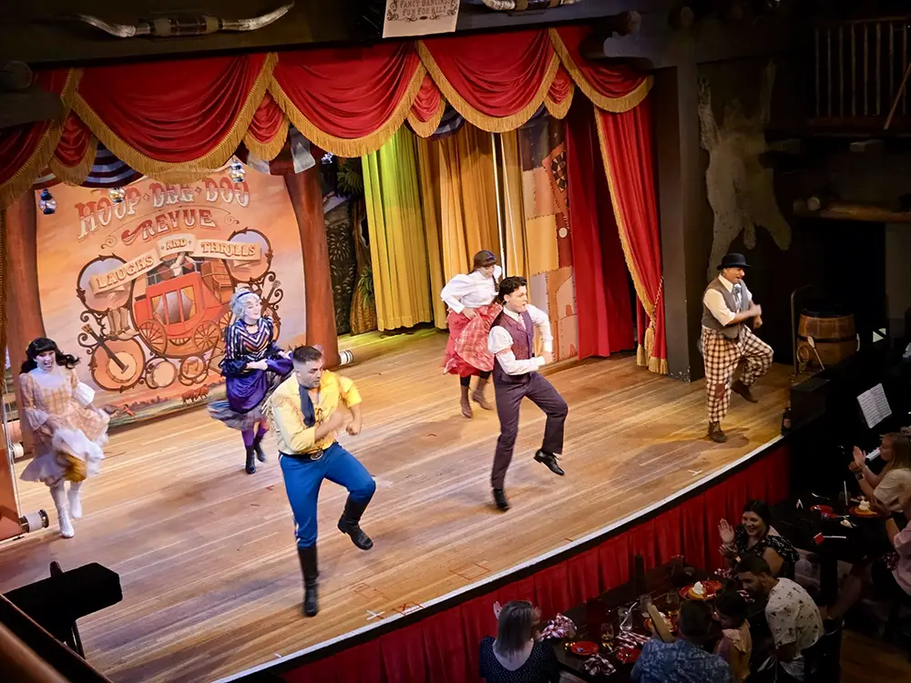 Five performers in historical frontier-style clothing dance on a wooden stage at Disney’s Fort Wilderness Resort, set against a red-curtained backdrop with an audience seated at tables in the foreground. - Favorite Grampy Travels