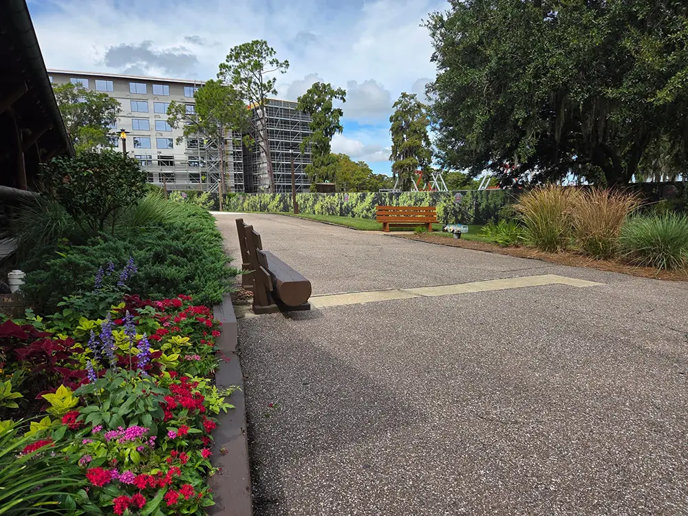 A paved path through a Disney Fort Wilderness garden with flower beds, two wooden benches, and a building under construction in the distance. - Favorite Grampy Travels