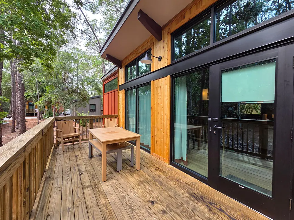 A wooden deck outside a modern cabin at Disney’s Fort Wilderness, featuring a dining table with two benches and a separate chair, alongside large glass sliding doors. - Favorite Grampy Travels