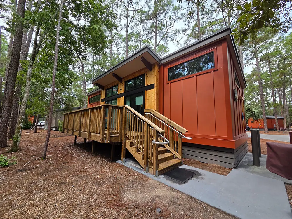 A modern small cabin with orange siding and a wooden deck and stairs surrounded by pine trees at Disney’s Fort Wilderness Resort. - Favorite Grampy Travels