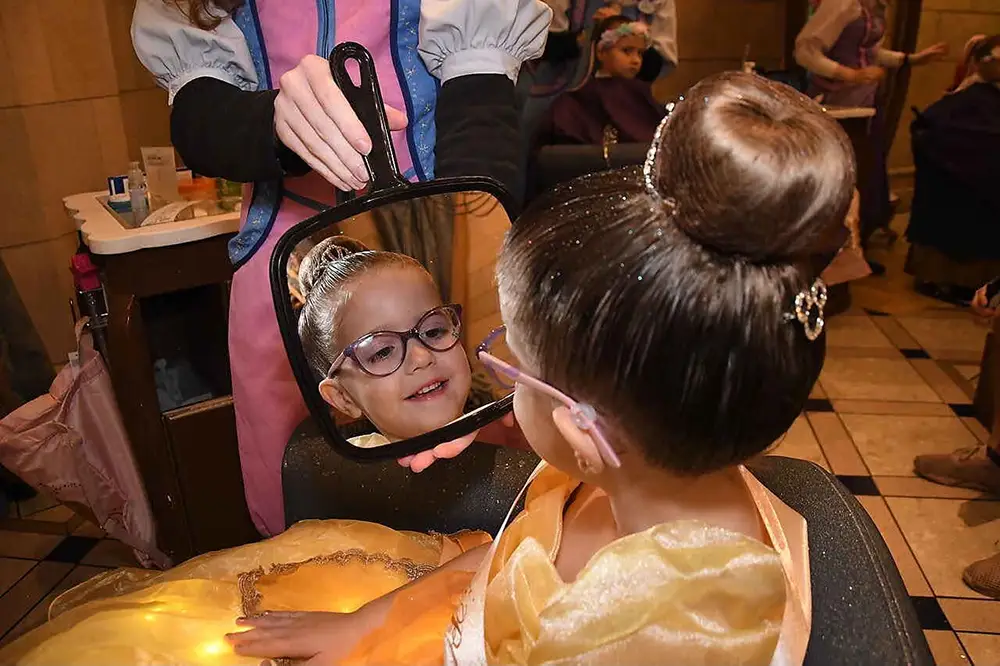 A young girl in a yellow princess dress looks into a hand mirror during her Bibbidi Bobbidi Boutique makeover, revealing her reflection as an attendant holds the glass. - Favorite Grampy Travels
