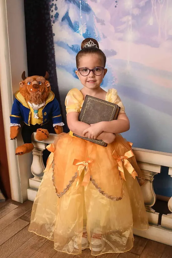 A young girl in a yellow Belle costume and glasses holds a book following her Bibbidi Bobbidi Boutique princess makeover, with a Beast stuffed animal sitting on a railing behind her. - Favorite Grampy Travels