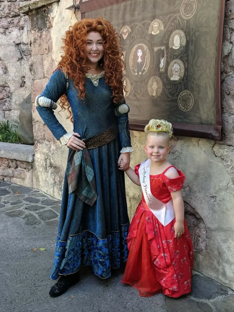 A woman dressed as Merida from Brave holds hands with a young girl in a red dress and a Birthday Princess sash after a Bibbidi Bobbidi Boutique makeover. - Favorite Grampy Travels