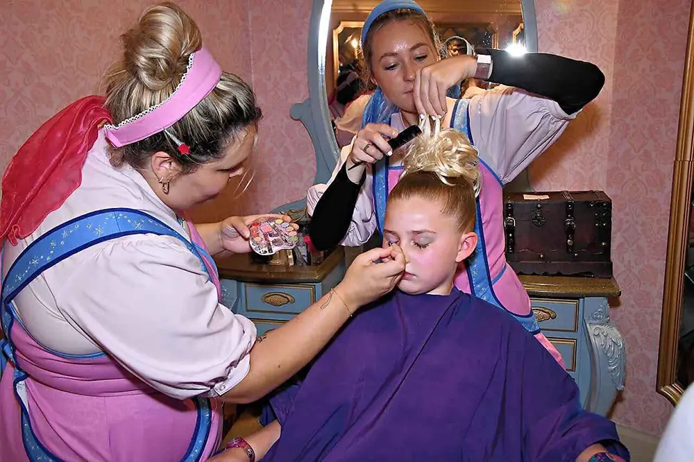 Two stylists in costume apply makeup and style the hair of a young girl sitting in a salon chair during her Bibbidi Bobbidi Boutique princess makeover. - Favorite Grampy Travels