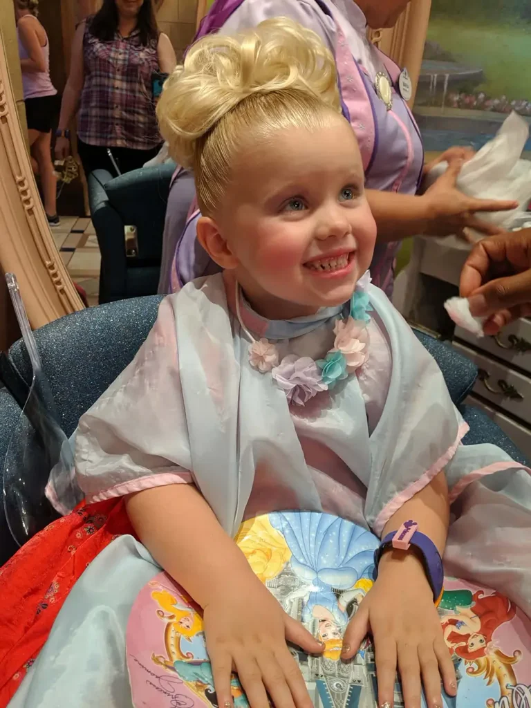 A young girl with a blonde updo sits in a salon chair wearing a light blue cape and floral necklace, smiling as she enjoys her Disney princess makeover at the Bibbidi Bobbidi Boutique. - Favorite Grampy Travels