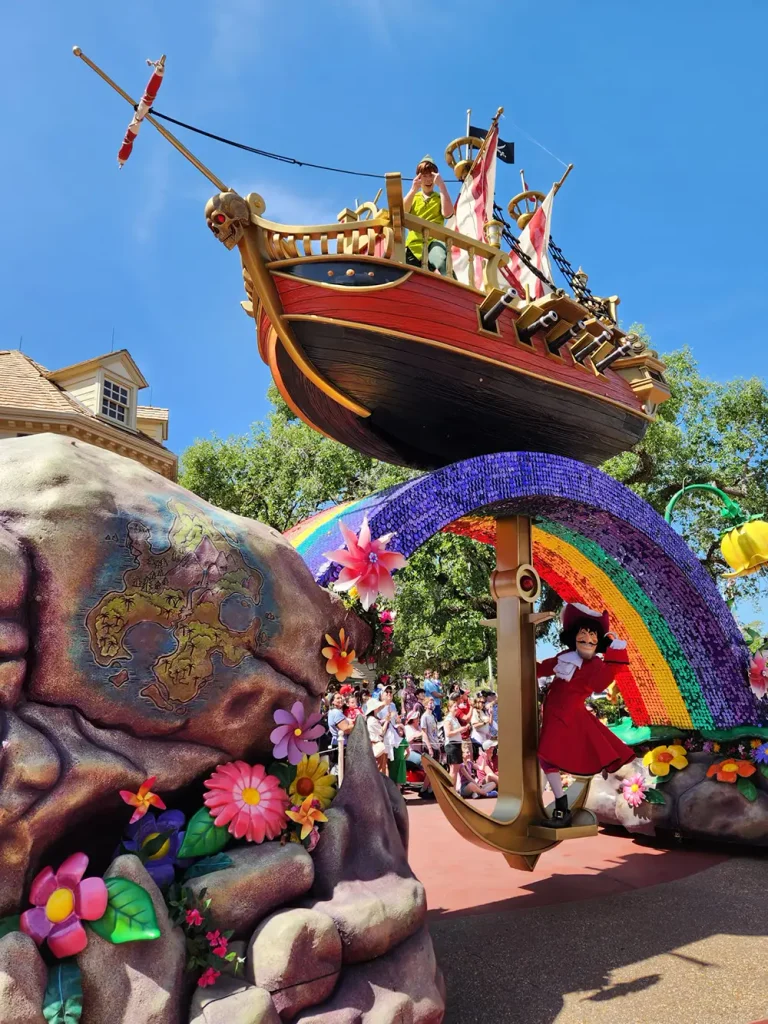 Peter Pan stands on the deck of a pirate ship float during Disney parades, while Captain Hook stands on a large anchor underneath a rainbow arch. - Favorite Grampy Travels