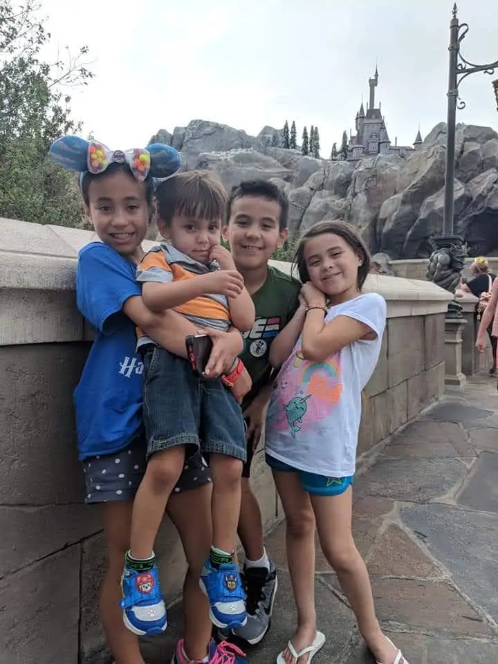 Wdw-magic-kingdom-grandkids-in-front-of-beasts-castle-be-our-guest - Favorite Grampy Travels Four smiling children posing together on a stone railing at a theme park with a castle in the background.