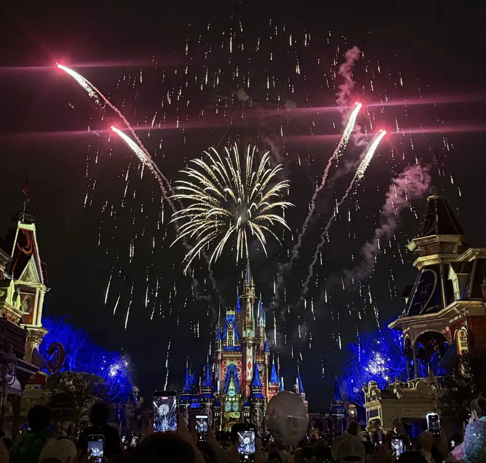 A crowd of people holds up their phones to watch Disney fireworks exploding over Cinderella Castle at night. - Favorite Grampy Travels