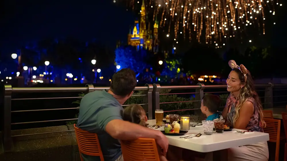 A family sits at an outdoor table at night to watch Disney fireworks explode over a distant, illuminated castle. - Favorite Grampy Travels