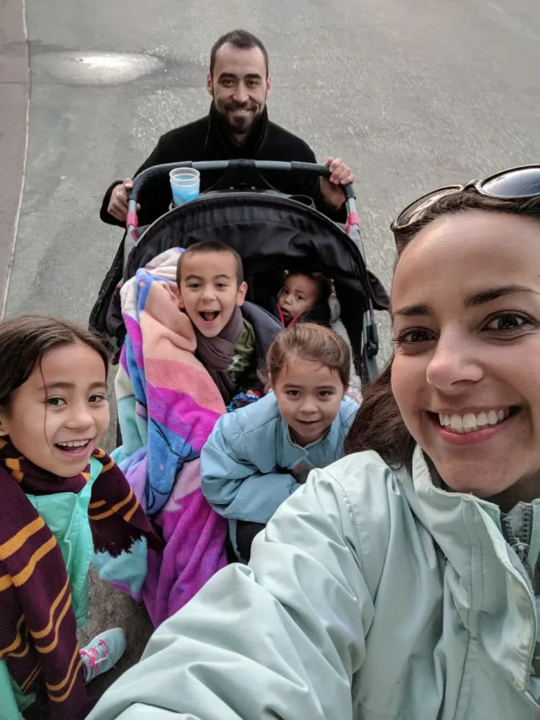 Wdw-nikki-sam-and-grandkids-selfie-with-stroller - Favorite Grampy Travels Smiling woman in the foreground takes a selfie as a man pushes a stroller with several children wrapped in colorful blankets behind her.