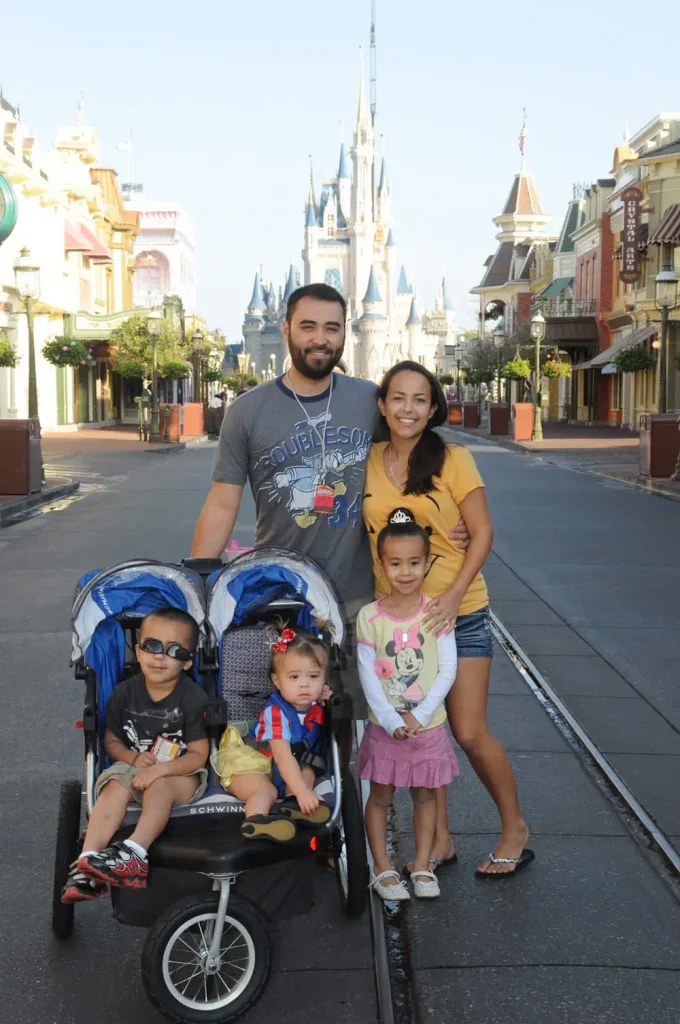 Wdw-nikki-sam-and-grandkids-at-magic-kingdom-with-stroller-castle-picture - Favorite Grampy Travels A family of five posing on aDisney park street with Cinderella Castle in the background; two kids sit in a double stroller while two others stand beside them.