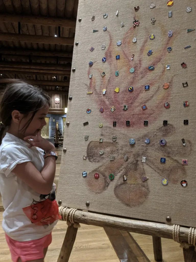 A young girl looks at a large burlap display board featuring a painted campfire with numerous collectible Disney pins attached to it. - Favorite Grampy Travels