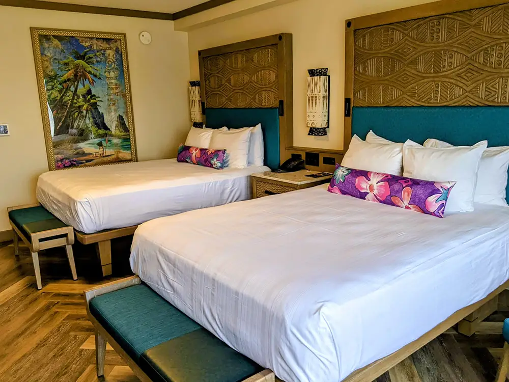 Wdw-polnesian-village-resort-room-double-queen - Favorite Grampy Travels Hotel room with two queen beds, teal headboards, white linens, and tropical accent pillows; wooden floor and framed beach painting on the wall.