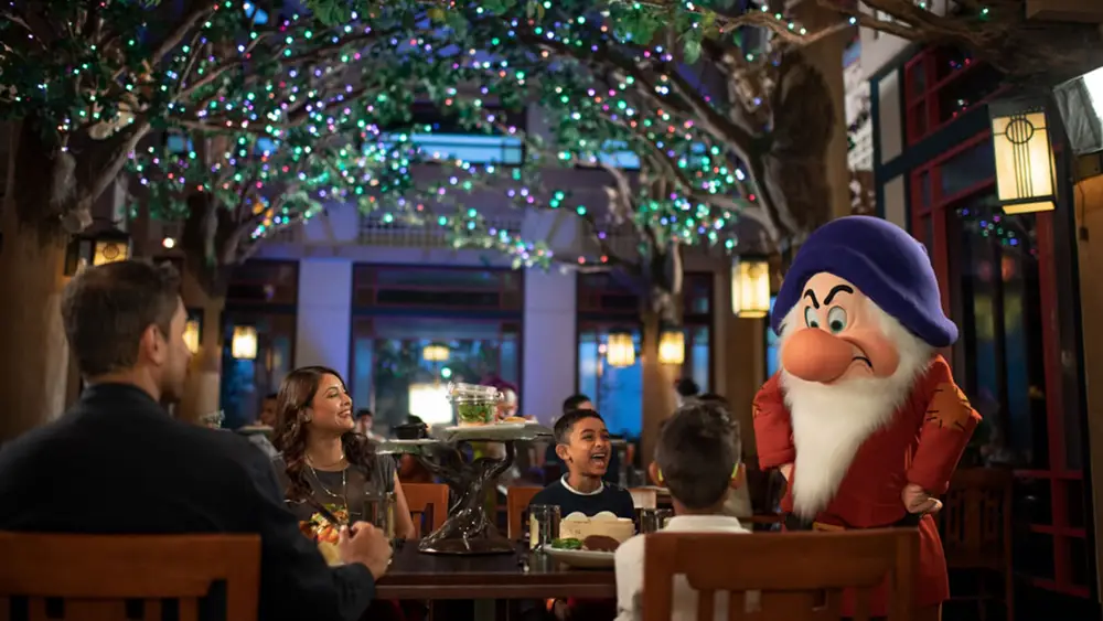 Grumpy the dwarf stands at a table in a dining room decorated with twinkling fairy lights, interacting with a family during one of the best Disney World dining character meals. - Favorite Grampy Travels