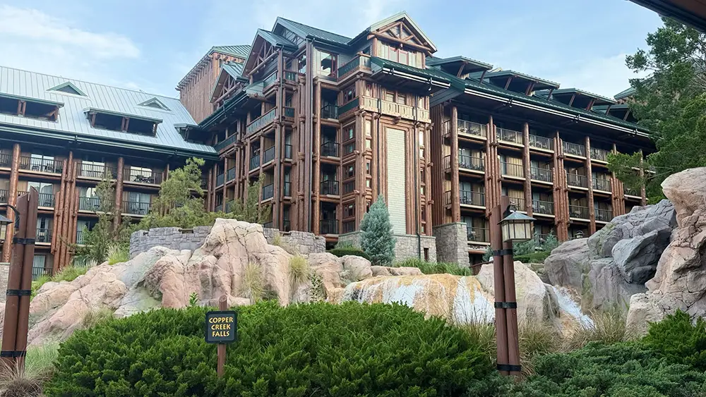 A large timber-framed building at Disney’s Wilderness Lodge nestled behind rock formations and a waterfall, with a Copper Creek Falls sign in the foreground. - Favorite Grampy Travels