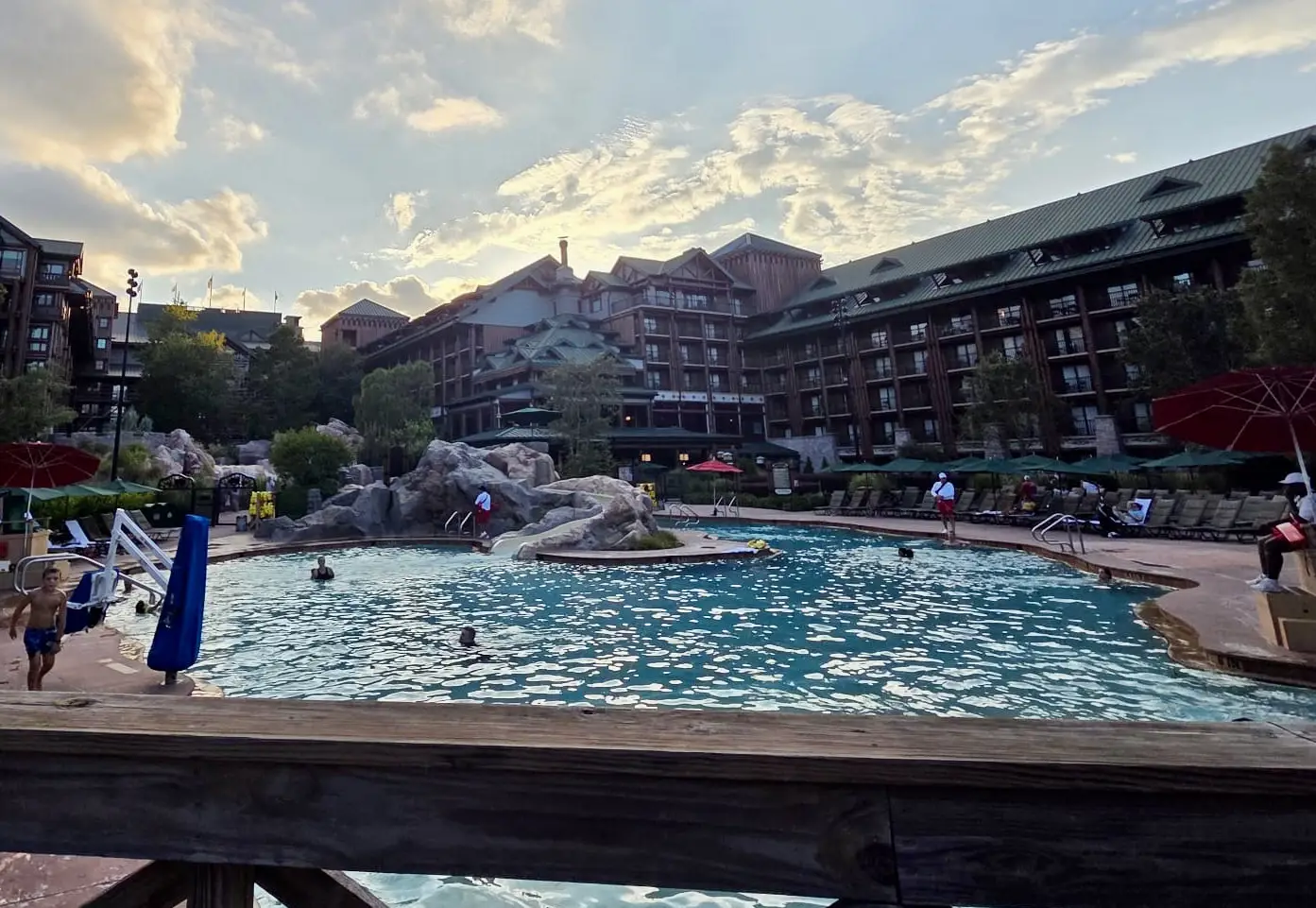 A large outdoor swimming pool at Disney’s Wilderness Lodge, featuring rocky landscaping and guests swimming, viewed from a wooden deck. - Favorite Grampy Travels