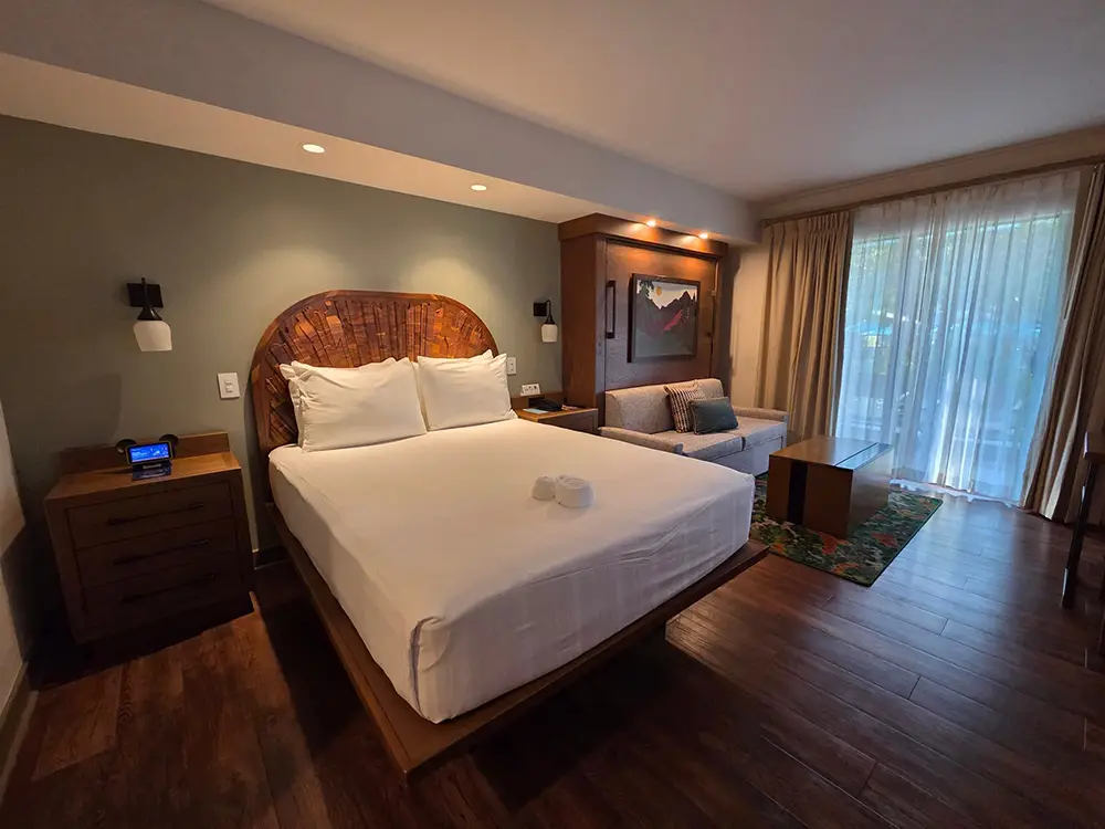 A hotel room at Disney’s Wilderness Lodge featuring a queen bed, a small sofa, and floor-to-ceiling curtains covering a balcony door. - Favorite Grampy Travels