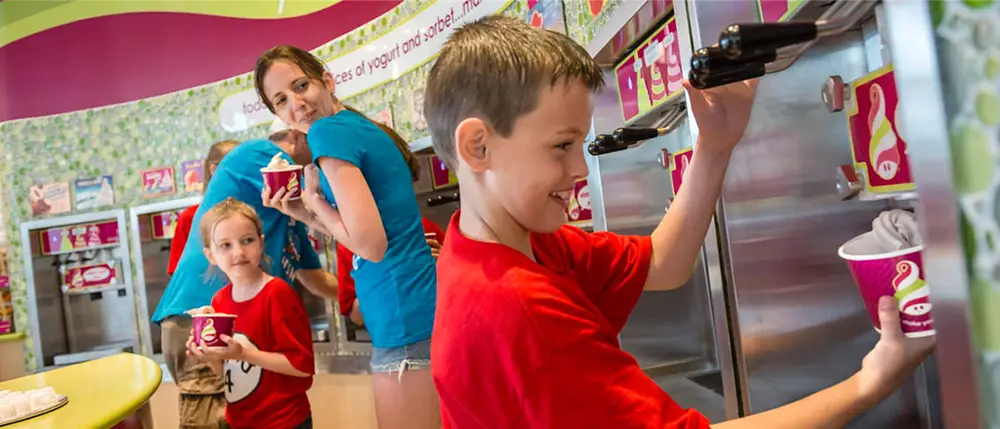 A boy in a red shirt dispenses frozen yogurt into a cup at a Universal CityWalk Orlando self-serve shop, while a woman and a young girl stand nearby holding their own cups. - Favorite Grampy Travels